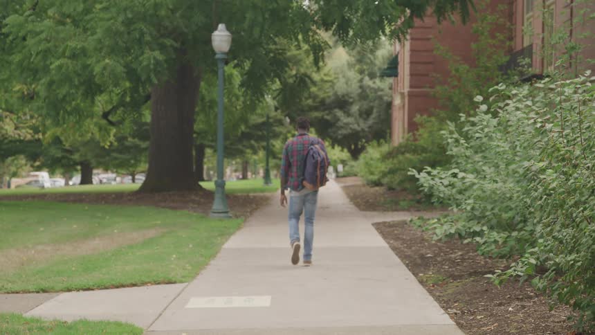 The video features a man with dreadlocks walking away from the camera down a sidewalk on a college campus he is wearing a plaid shirt and carrying a backpack suggesting he is a student heading to or from classes