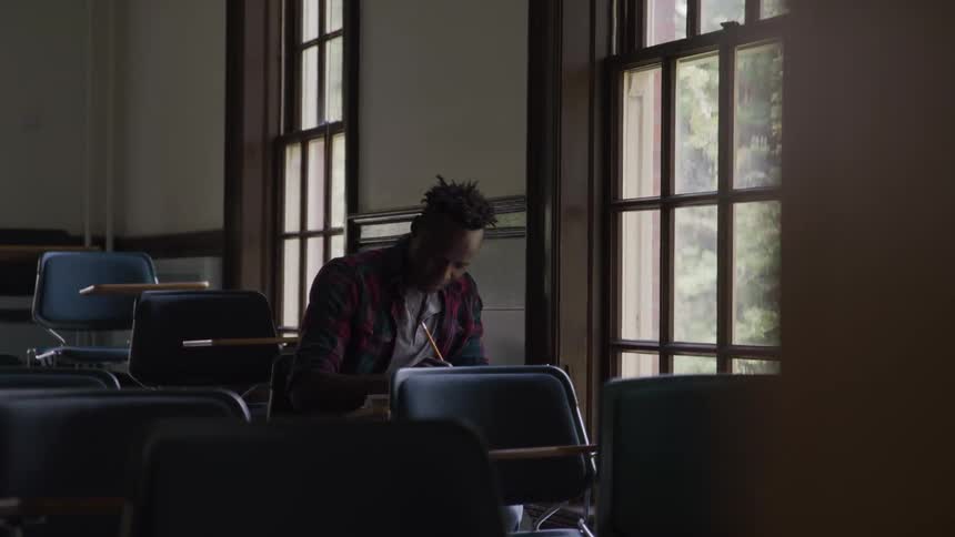 The video shows a male student with dreadlocks sitting at a desk in a classroom and diligently writing in a notebook or textbook with a pencil the setting appears to be a classroom with rows of desks and large windows in the background