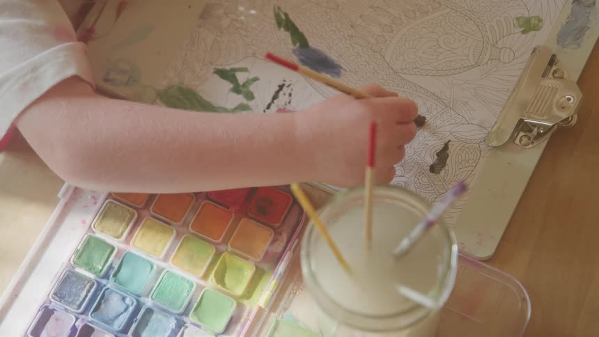 The video captures an overhead shot of a persons hands diligently coloring in a coloring book with a paintbrush showcasing the detailed and vibrant application of colors a watercolor palette is visible in the bottom left alongside a glass of murky wat