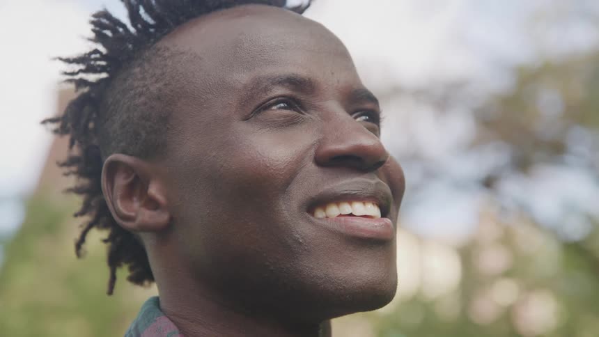 The video features a man with dreadlocks smiling and looking towards the upper right of the frame with a bright natural light illuminating his face the background is softly blurred showing hints of green foliage and a clear sky suggesting an outdoor