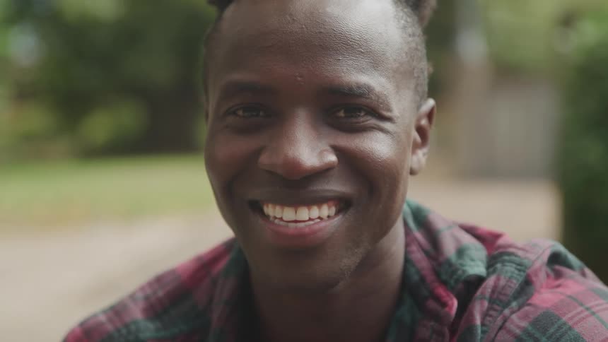 The video features a man with dreadlocks looking directly at the camera with a wide genuine smile he is wearing a red and green plaid shirt and the background is blurred with hints of green and concrete suggesting an outdoor setting