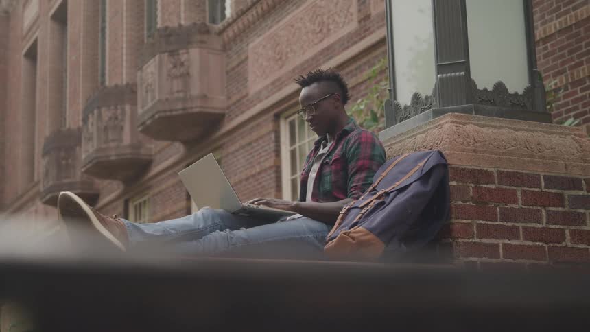 A man is sitting outside typing on his laptop
