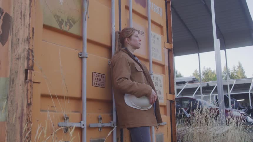 The video shows a woman with reddishblonde hair leaning against an orange shipping container holding a white hard hat she is looking towards the left side of the frame with a serious expression and other industrial structures are visible in the blurre