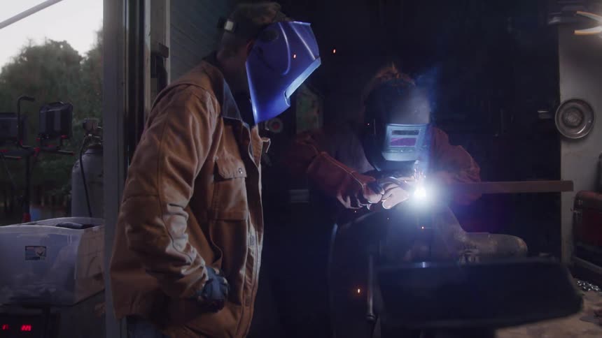 The video shows two individuals engaged in welding outdoors both wearing protective helmets one welder is actively joining metal with a bright arc while the other stands nearby observing