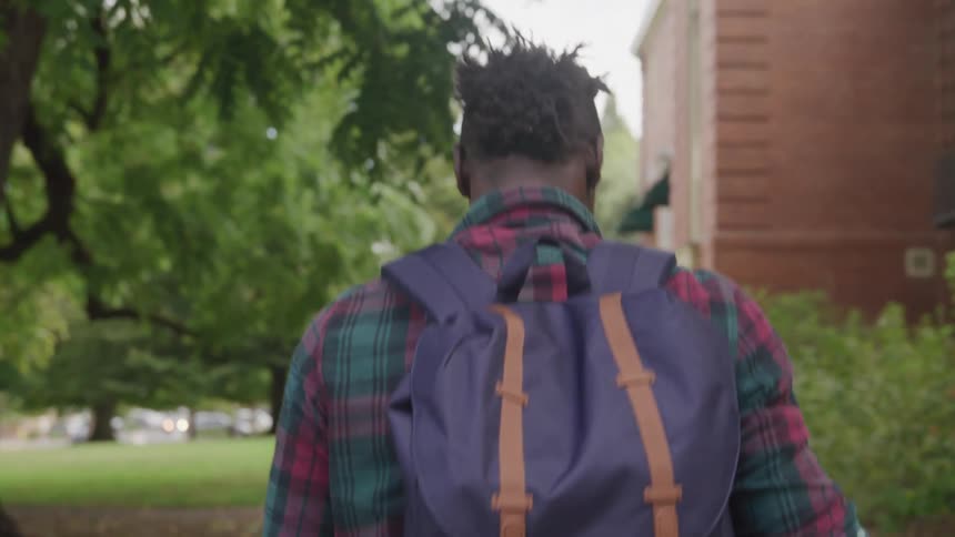 A man wearing a backpack walking on a sidewalk with trees on one side and a brick building on the other