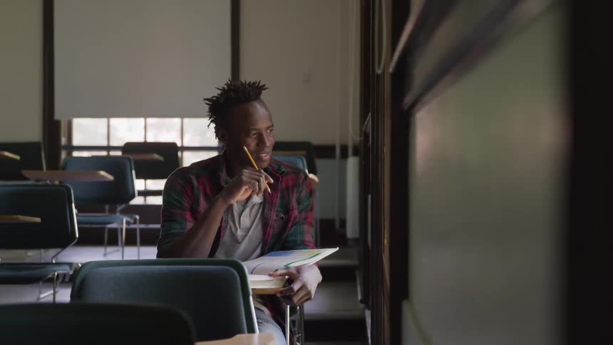 The video features a male student with dreadlocks looking thoughtfully towards the upper right of the frame while holding a pencil to his chin in a classroom setting empty desks and chairs are visible around him suggesting he might be alone in the room