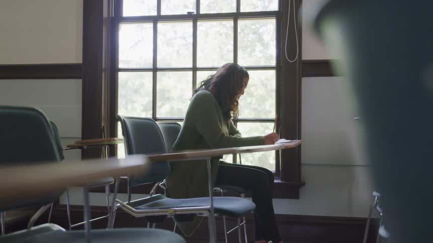 The video features a female student with long dark hair wearing a green cardigan and dark pants seated at a desk near a large window she is diligently writing in a notebook or on a piece of paper suggesting an academic or study setting