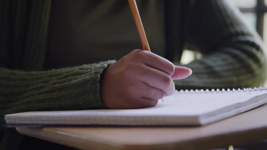 The video shows a closeup of a persons hands holding a pencil and writing diligently in a notebook the hand wears a subtle ring on the ring finger and the background is blurred suggesting a focused work or study environment
