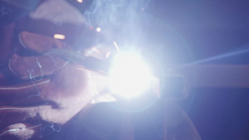 A person wearing welding gloves uses a welding torch to create sparks and bright light while working on a metal structure