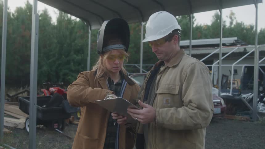Two construction workers one wearing a welding helmet and the other a hard hat review a clipboard together under a metal canopy at a work site surrounded by trees and equipment
