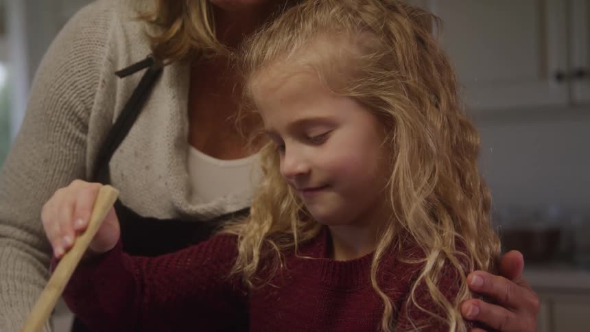 The video shows a child with long curly blonde hair standing next to an adult learning to stir a bowl with a wooden spoon the scene is likely in a kitchen indicating a shared activity possibly cooking or baking