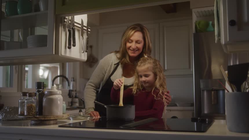 Girl holding a wooden spoon and cooking in a pot accompanied by her mother