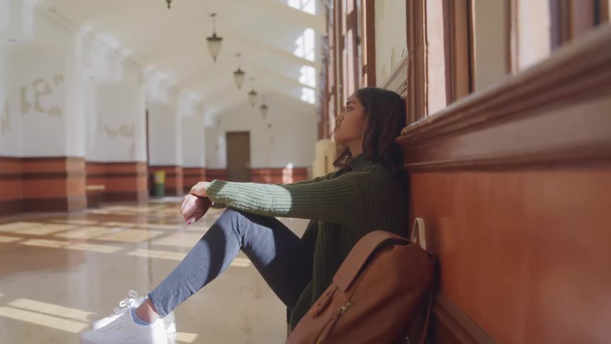 The video features a female student seated on the floor of a sunny hallway leaning against a wooden wall with a backpack beside her appearing deep in thought or troubled the sunlight casts long shadows across the polished floor highlighting the archit