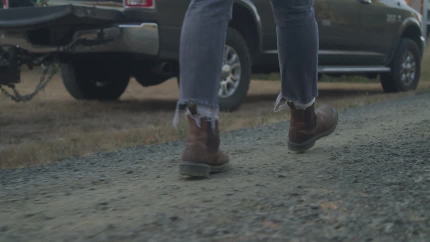 The video shows a person from the waist down walking away from the camera on a gravel path wearing blue jeans and brown boots a pickup truck with a trailer hitch is visible in the background suggesting a rural or industrial setting