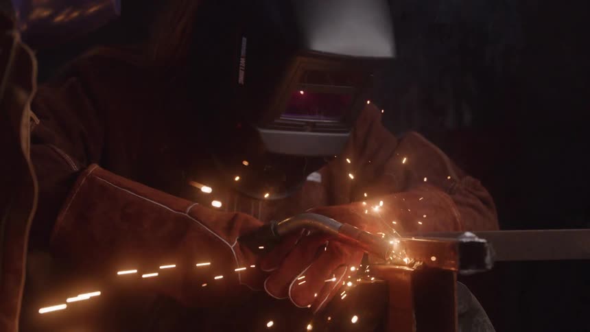 The video shows a welder wearing a protective helmet and heavy gloves actively working on a metal surface sparks fly brightly from the point of contact illuminating the dark workshop environment