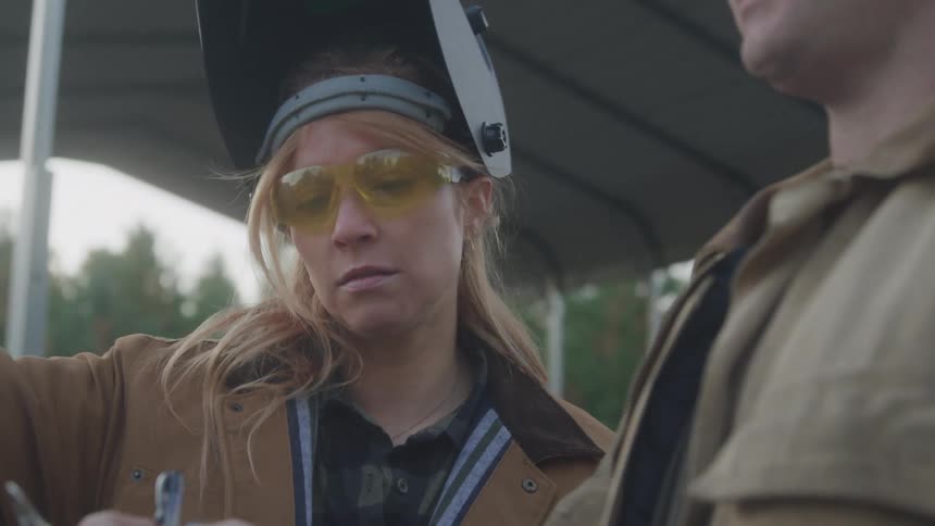 A woman wearing a welding helmet pushed back yellow safety glasses and a brown jacket talks to a man in a tan jacket standing next to her who is partially visible on the right side of the frame