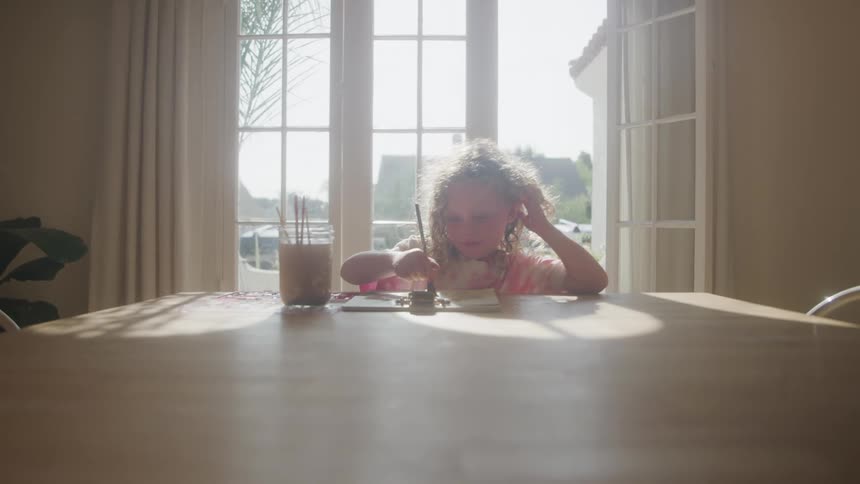 A young girl paints at a sunlit table softly backlit by large windows