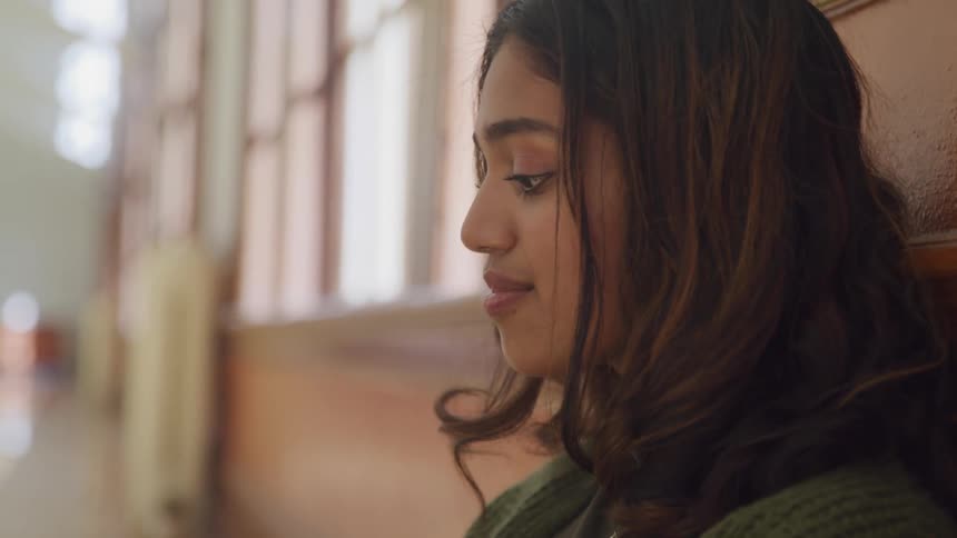 The video shows a young woman looking down with a pensive expression her long dark hair cascading over her shoulders she appears to be listening intently or reflecting on something with soft light illuminating her from the side