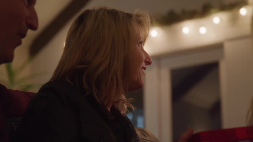 A blonde woman is sitting next to a man and she is smiling and looking forward to where a little girl wearing a red hat appears