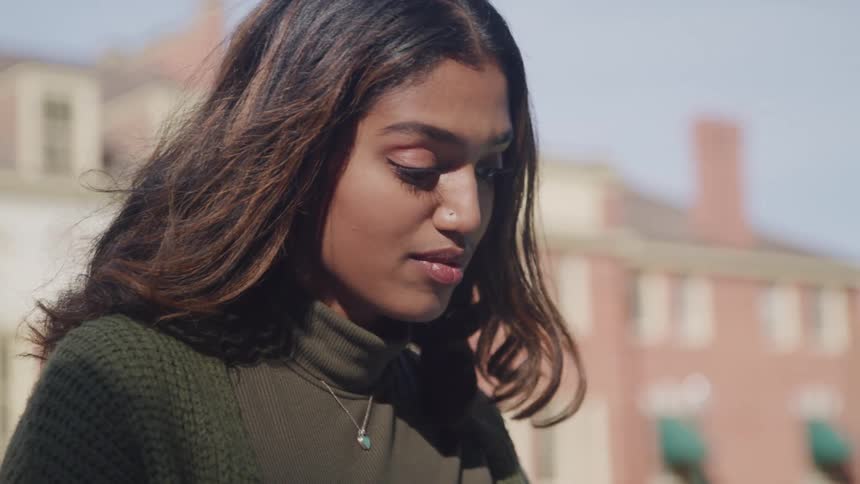 Woman in a knit sweater smiles as she thinks in front of large school buildings