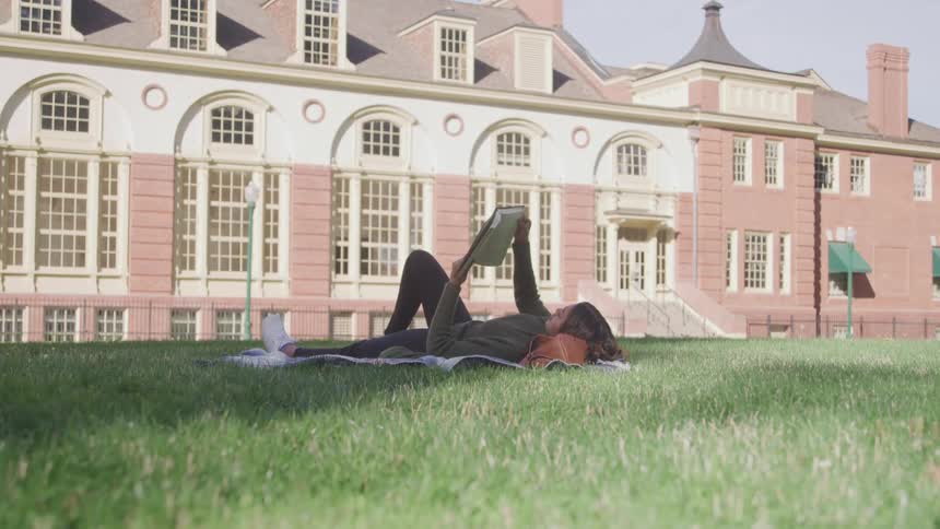 The video shows a student lying on their back on a grassy lawn in front of a large traditional university building reading a textbook the architecture of the building with its many windows suggests a classic academic setting