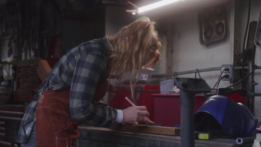 The video shows a person with long blonde hair and a plaid shirt working in a dimly lit workshop appearing to be engaged in a crafting or woodworking project tools and equipment are visible in the background suggesting a creative and handson environm