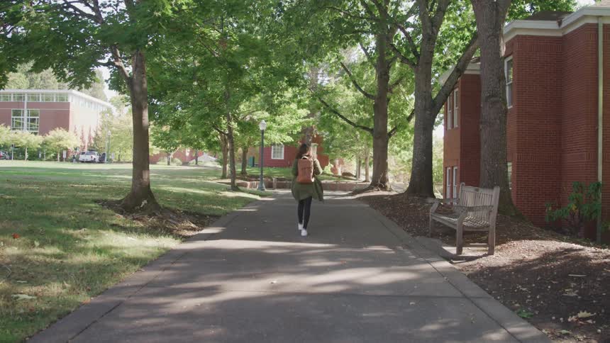 The video features a female student walking away from the camera down a treelined path on a college campus wearing a green cardigan and carrying a brown backpack large academic buildings are visible on either side of the path emphasizing the universit