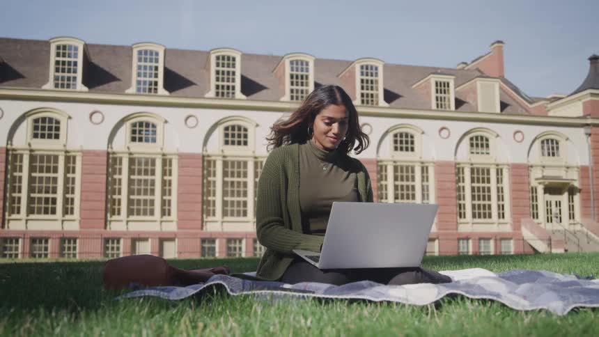 Woman sitting on the grass typing on a laptop with a building in the background