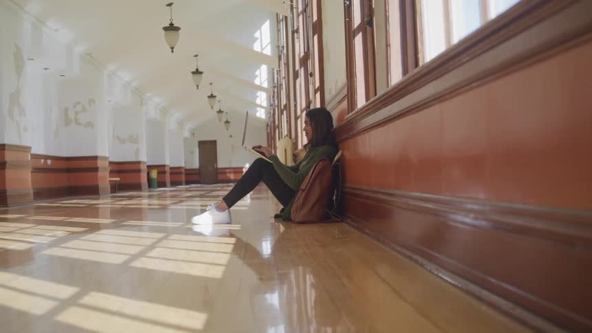 Woman sitting in the hallway of a building typing on a laptop