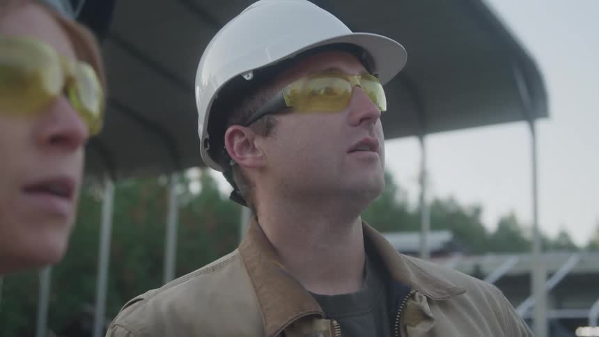 Two workers wearing hard hats and safety glasses discuss their work while onsite