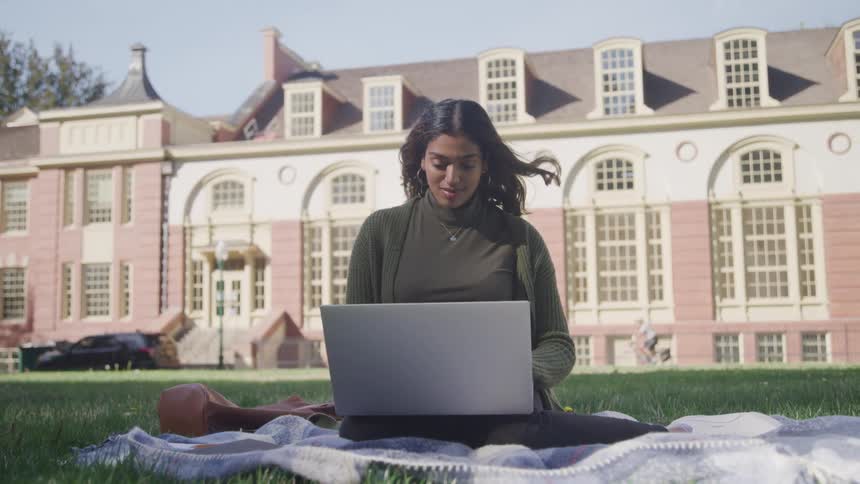 Woman sitting on a cloth in the grass of a university campus typing on a laptop
