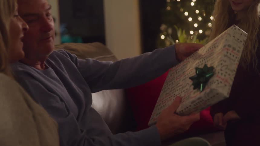 Little girl handing a gift to her father who is sitting on the couch next to her mother at christmas time