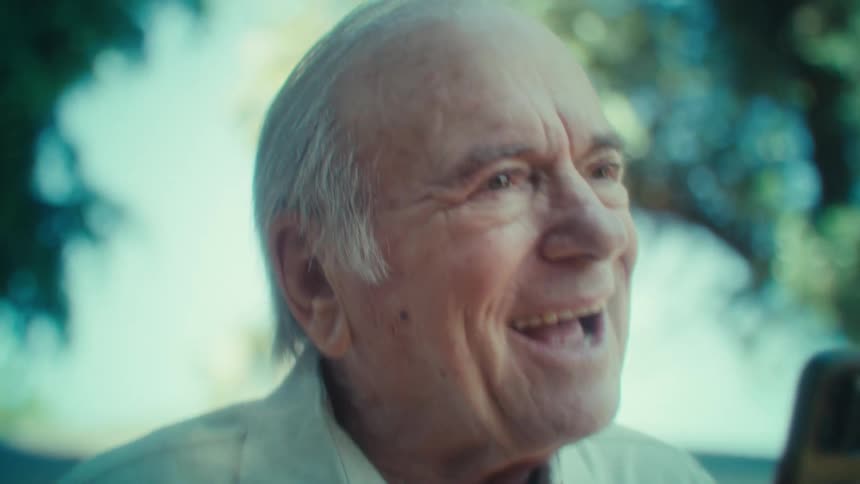 Closeup of an older caucasian man wearing a beige collared on an outdoor setting with natural light with trees in the back
