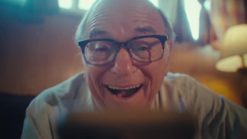 Elderly man wearing glasses and a white shortsleeved collared shirt laughing to a smartphone screen