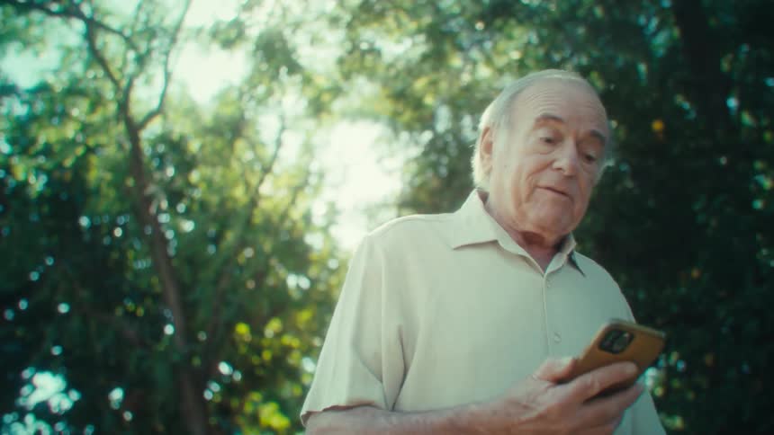 The video shows a closeup of an elderly man with short white hair and a lightcolored polo shirt smiling softly while holding a phone the background features lush green foliage suggesting an outdoor setting