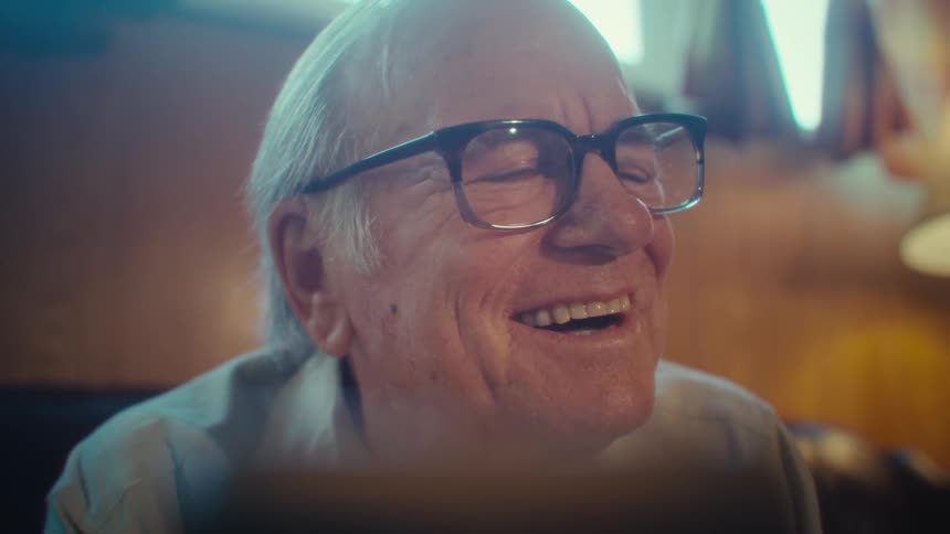 An elderly man is sitting in a room smiling at a tablet in front of him