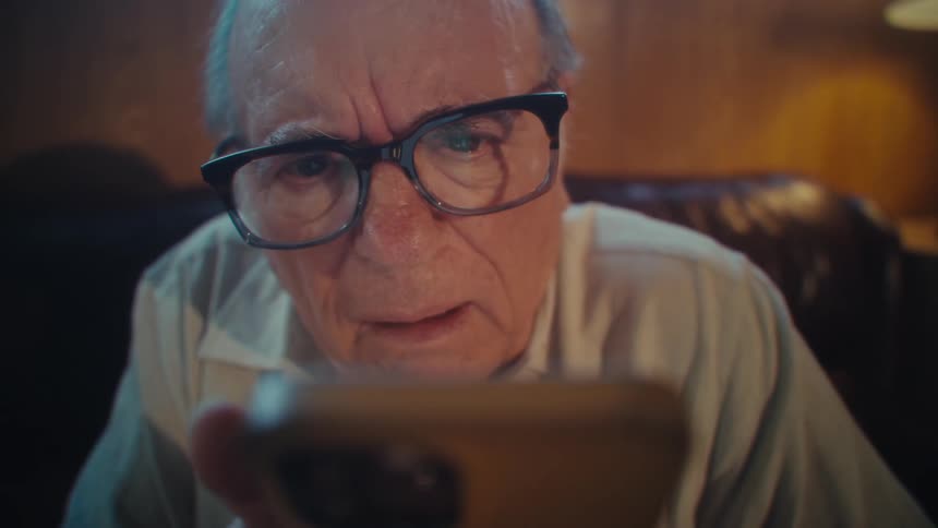 Elderly man sitting and looking surprised while interacting with a smartphone