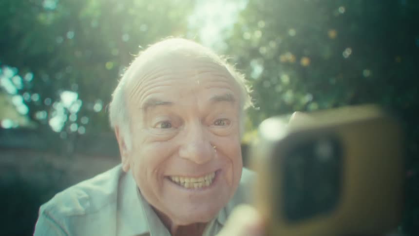 Elderly man vibrating and laughing to a smartphone with trees in the back