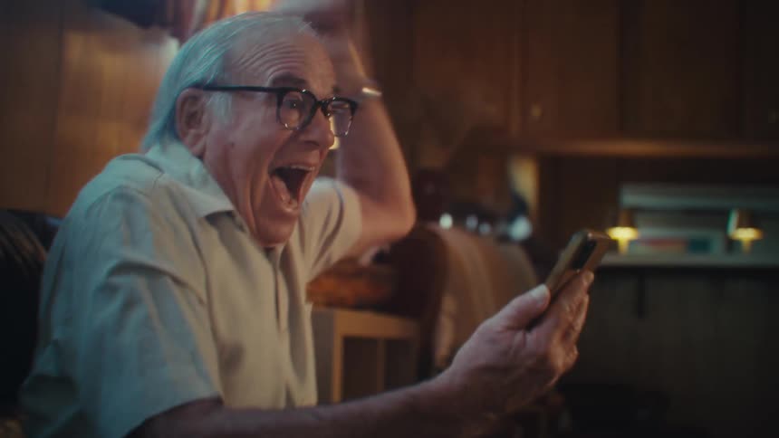 Elderly man exclaims and celebrates while watching media content on a phone in a living room