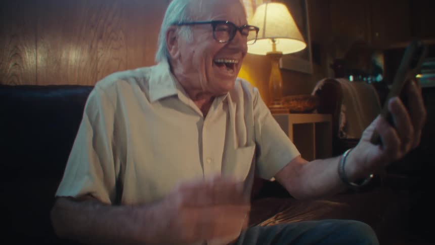 Elderly man laughs and celebrates while watching media content while sitting in a living room during daytime