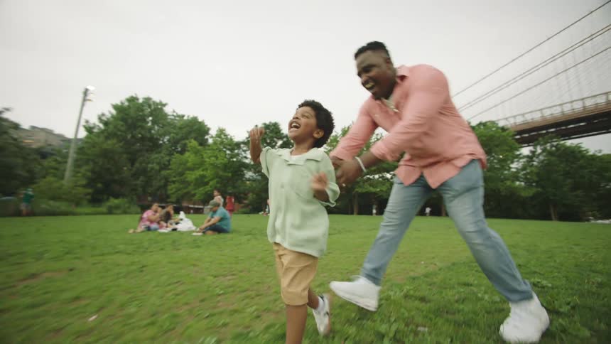 Father and son running and playing in an outdoor park with other people sitting on the grass