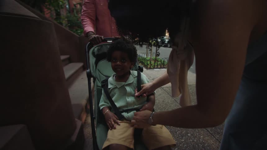 Mother arranges and straps her son in a child stroller on a walkway outdoors during daytime