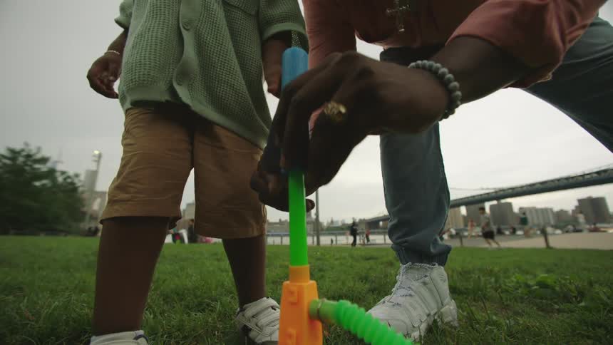 A child stands on grass beside a colorful toy rocket launcher while an adult helps position the rocket with a bridge and city skyline visible in the background