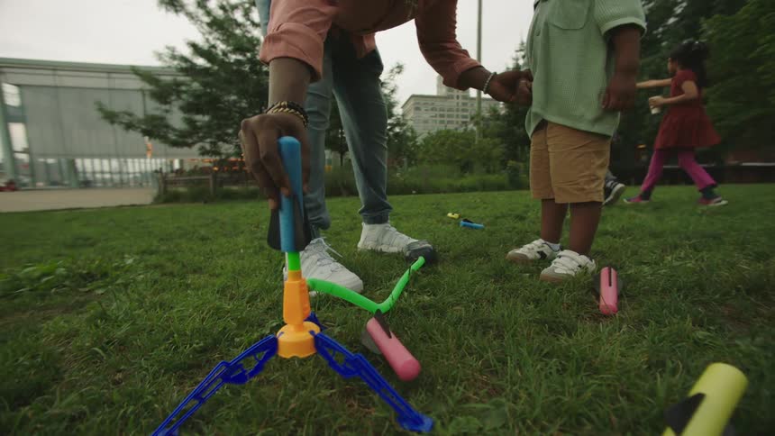 The video shows a lowangle shot of an adult and a young child in a parklike setting playing with bright green and blue toys that resemble a catch game with nets in the background a bridge is visible suggesting an urban park next to a body of water