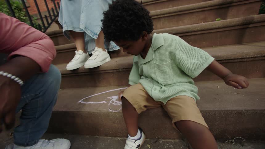 A young boy sits on brownstone steps drawing with sidewalk chalk while his smiling parents watch nearby creating a warm and playful family moment