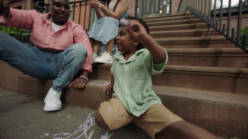 A black family are playing with their son who is drawing with chalk outside