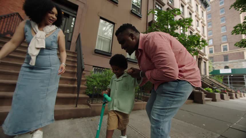 Family helps child ride scooter on a city sidewalk
