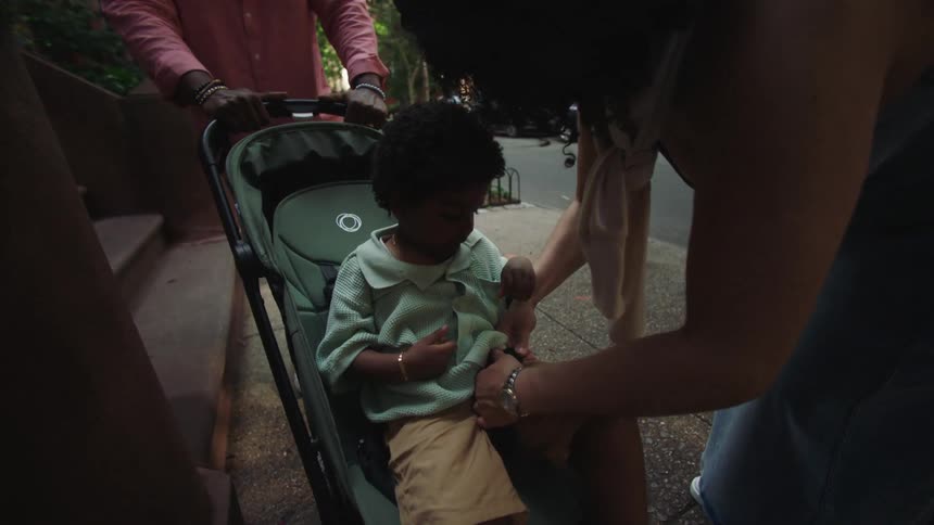 Father holds the stroller and mother belts their son into it while they stand on the sidewalk in a residential neighborhood on a summer day