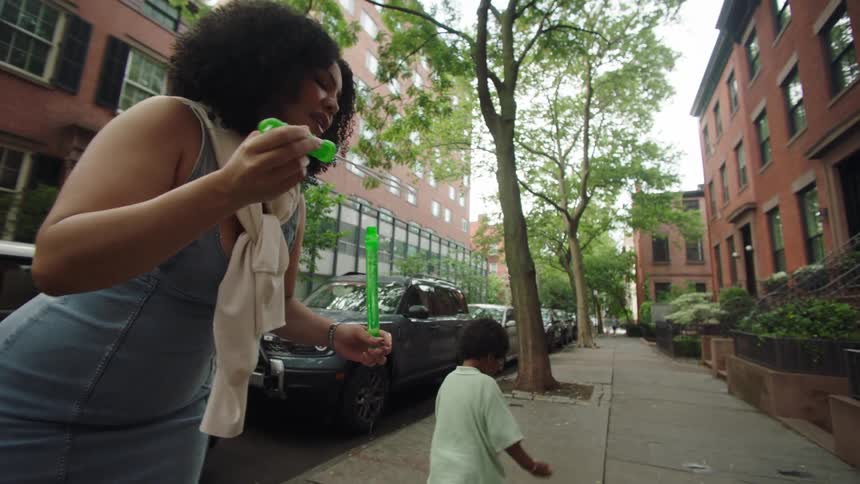 A man and a woman on a sidewalk playing with their child and the woman is blowing soap bubbles towards him