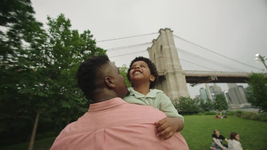 A man in a pink shirt carries a child in a park beneath a bridge with people relaxing on the grass with a city skyline in the distance